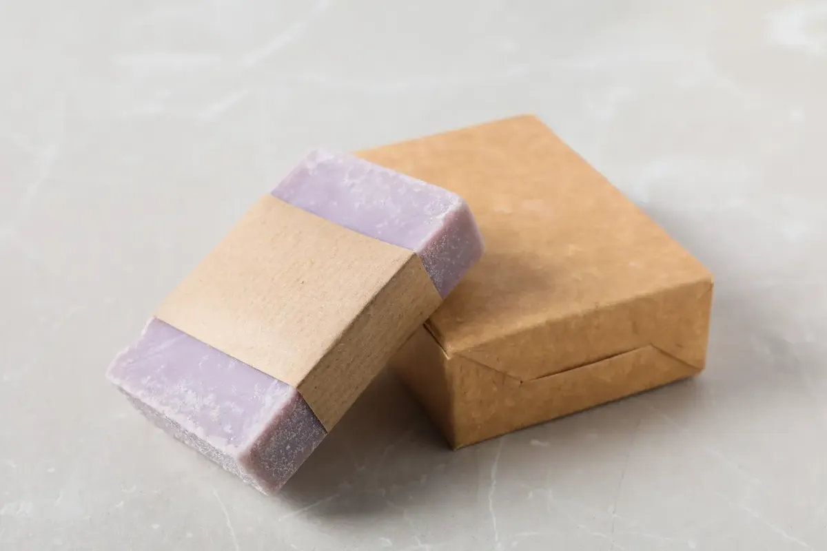 Un sapone solido artigianale avvolto in carta riciclata accanto a una scatola di cartone, che evoca prodotti naturali e sostenibili