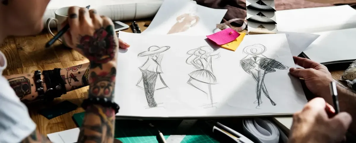  Mani tatuate di un designer che esamina schizzi di figurini di moda, simbolo della creatività nel design