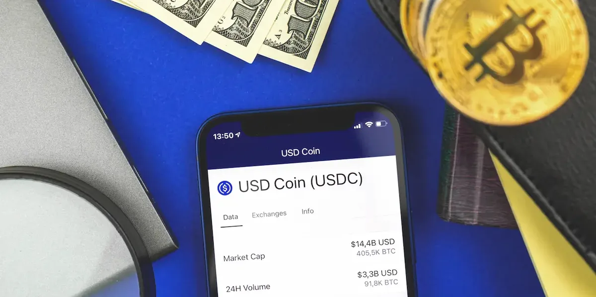 Schermo di smartphone che mostra valuta criptografica USD Coin, con banconote in dollari e Bitcoin, che simboleggia i pagamenti digitali