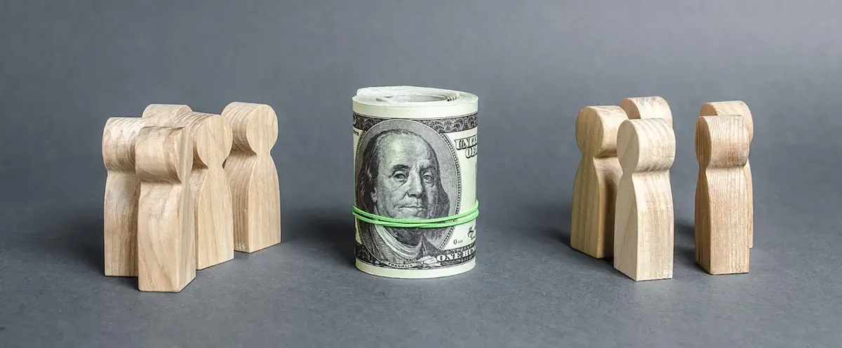 Rappresentazione concettuale del crowdfunding: un rotolo di banconote da cento dollari tra gruppi di statuette in legno, simboleggiando il capitale della folla