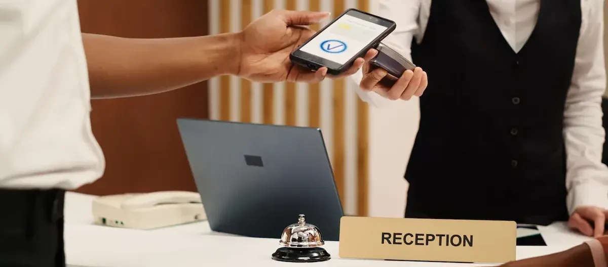 Un cliente che utilizza uno smartphone per interagire con un terminale POS alla reception di un hotel, illustrando il check-in digitale e i servizi smart