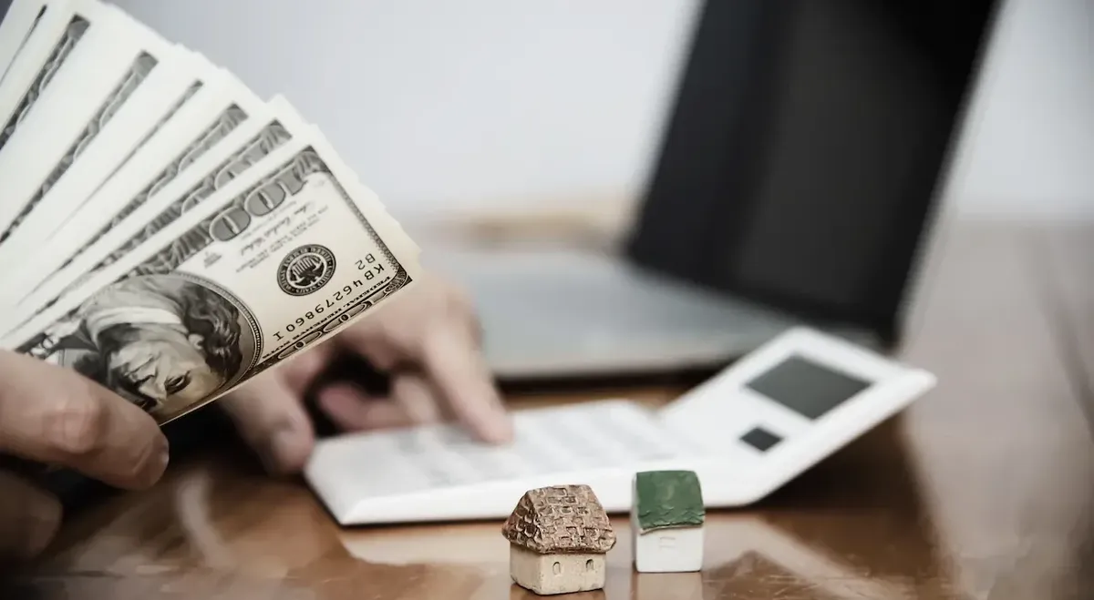 Mani che contano dollari con una calcolatrice e modellini di case, simboleggianti la pianificazione finanziaria e gli investimenti immobiliari negli Stati Uniti.