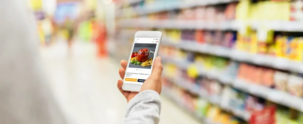 Persona in un supermercato che confronta prodotti fisici e online tramite smartphone, a simboleggiare lo shopping moderno e l'integrazione tecnologica.