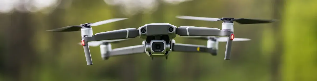 Un drone grigio in volo, con luci LED rosse e telecamera visibile, contro uno sfondo naturale sfocato