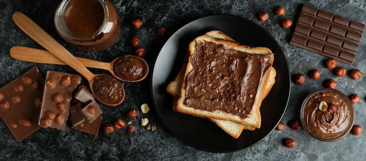 Pane tostato con crema spalmabile al cioccolato, nocciole sparse, vasetto di crema e tavolette di cioccolato su un tavolo scuro