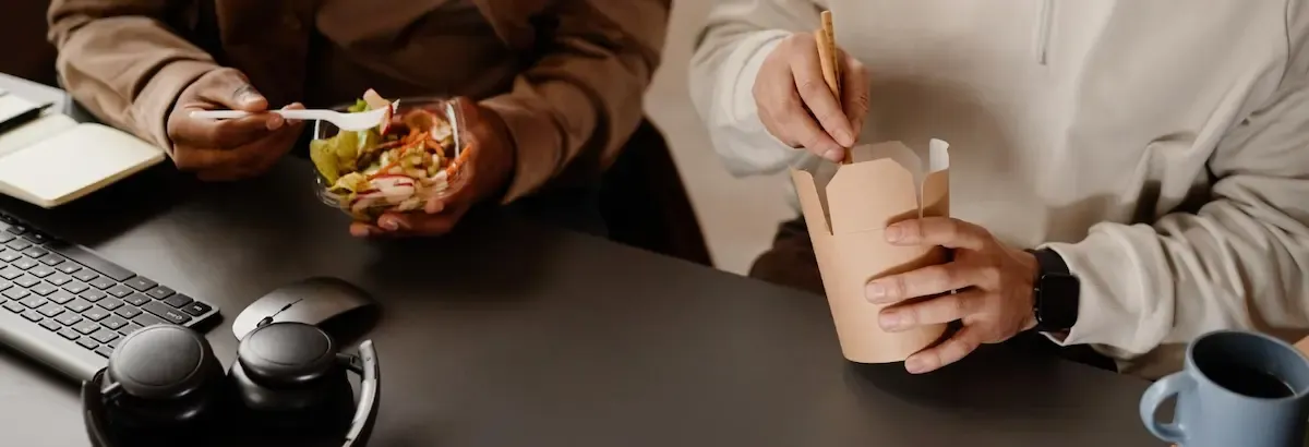 Due persone pranzano alla scrivania, una con insalata e l'altra con cibo take-away, in un ambiente d'ufficio
